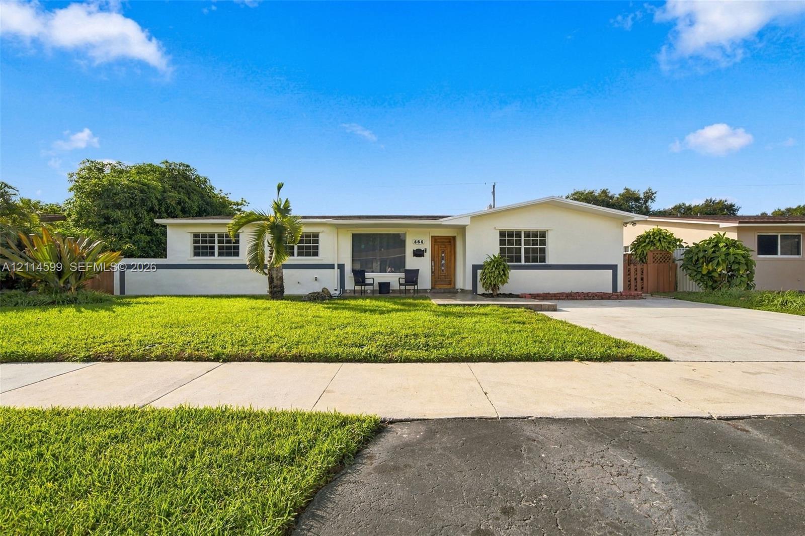 Single-story exterior featuring light-toned stucco siding, horizontal dark-toned accent trim, a concrete driveway, and a front lawn with established landscaping