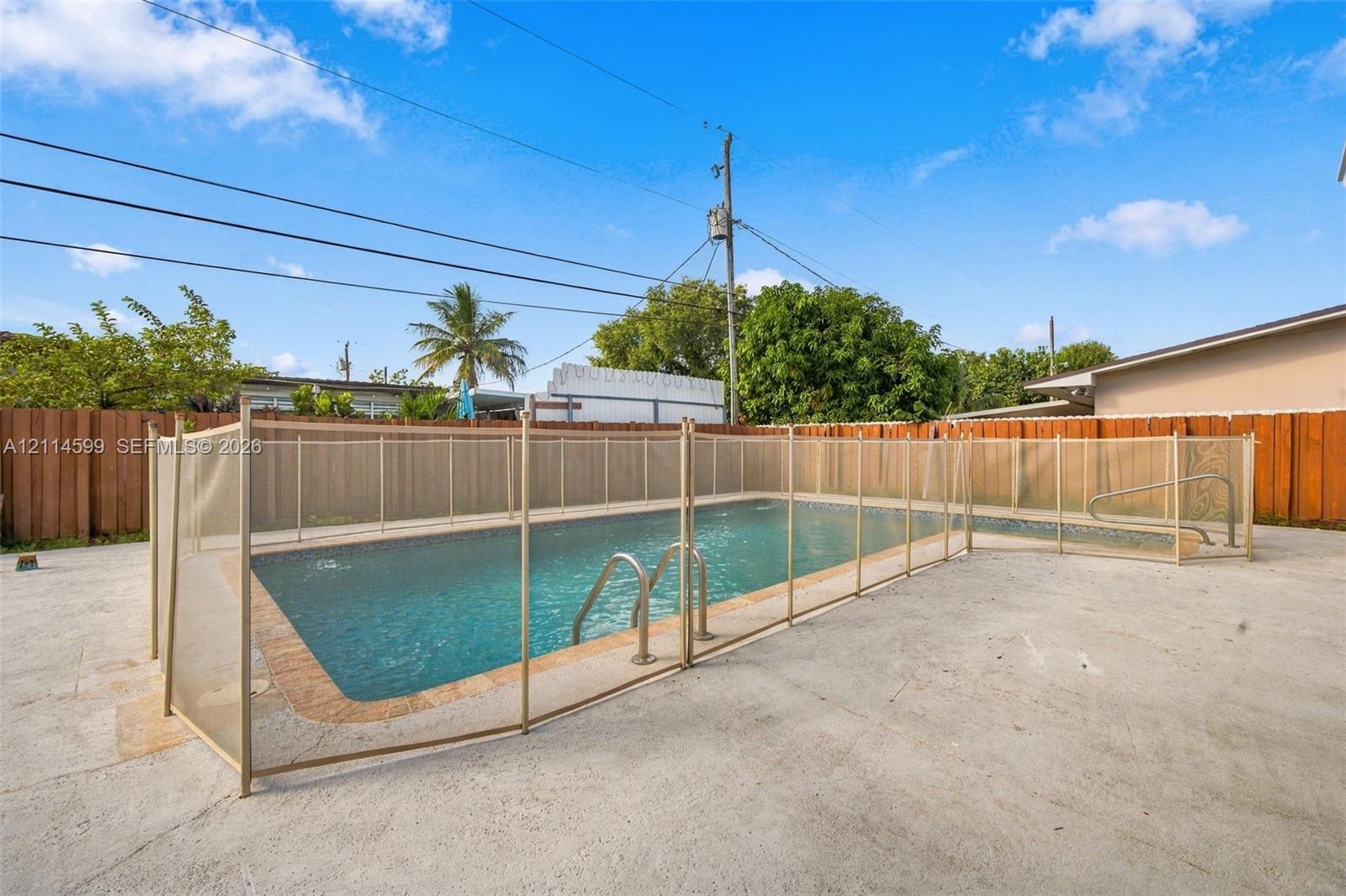 In-ground swimming pool with a stone-finish perimeter, metallic pool ladder, and encompassing mesh security fencing, set within a concrete patio enclosure and surrounded by a wood privacy fence