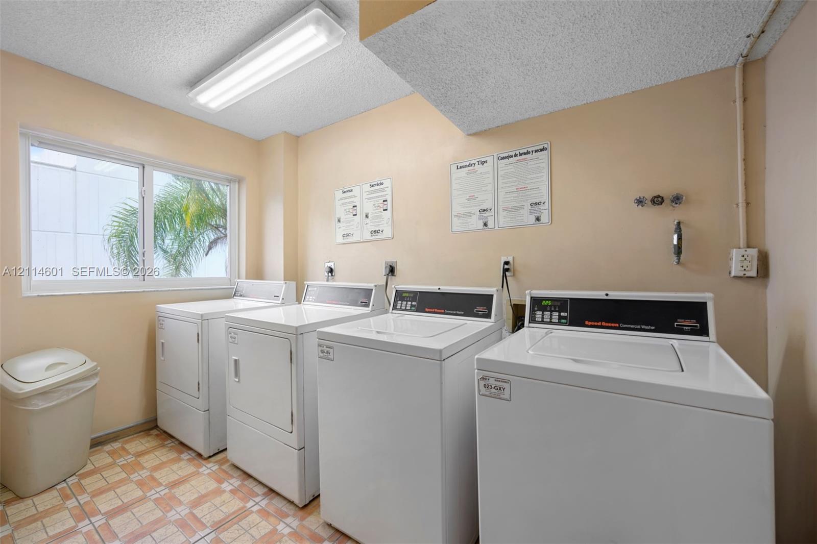 Laundry room featuring multiple high-capacity washer and dryer units, a large window, fluorescent overhead lighting, and durable tiled flooring