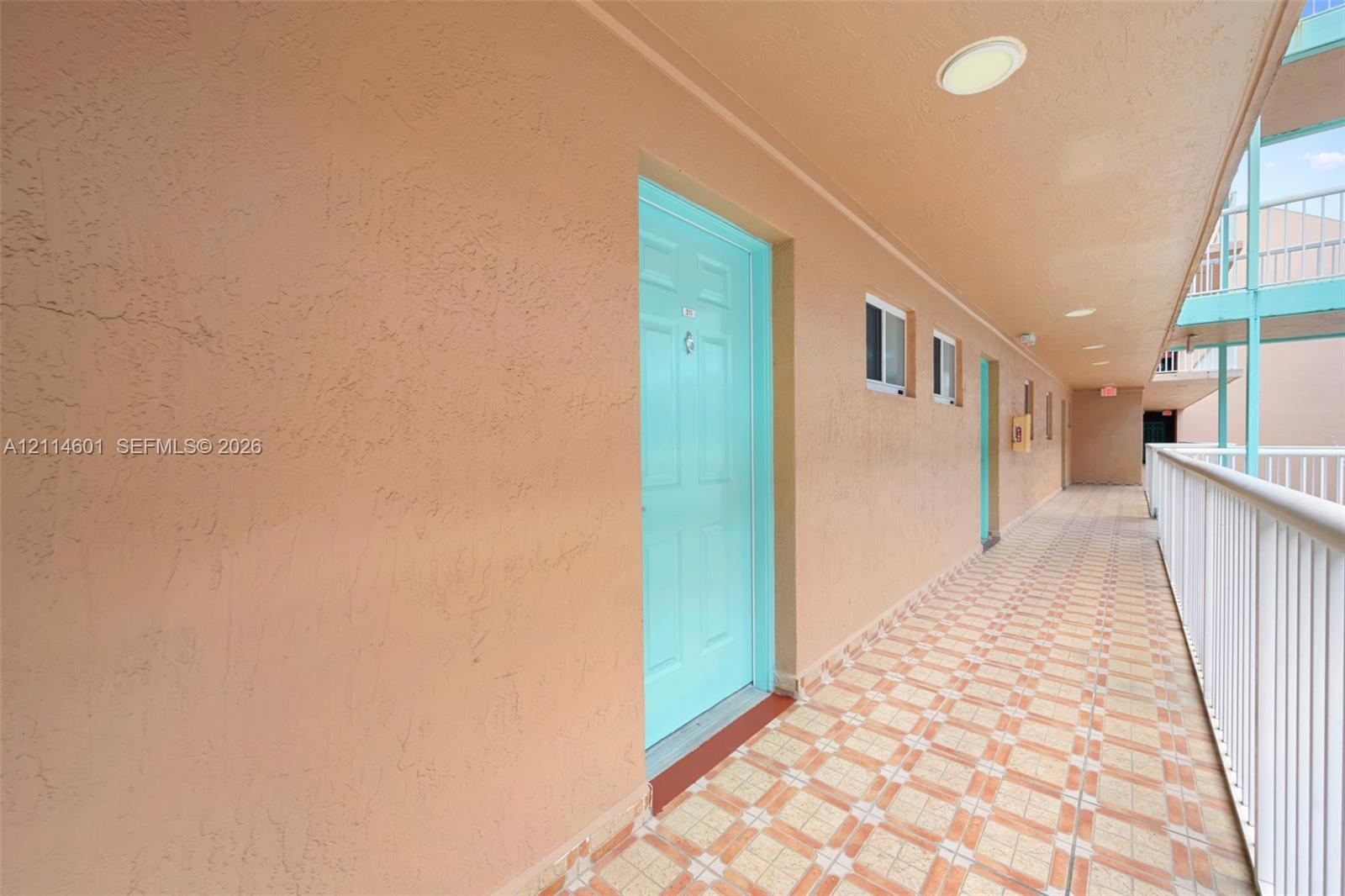 Exterior walkway featuring textured stucco walls, tiled flooring, recessed ceiling lighting, and white metal railing Exterior walkway featuring textured stucco walls, tiled flooring, recessed ceiling lighting, and white metal railing
