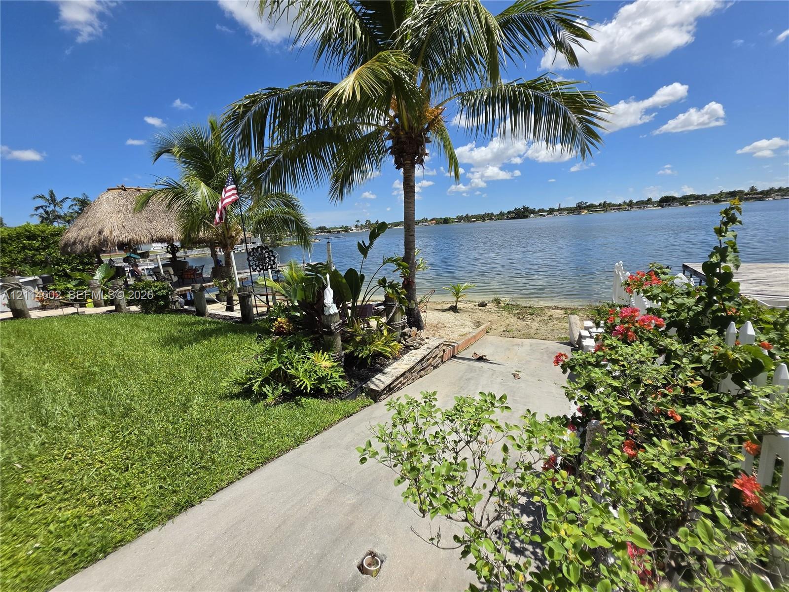 Waterfront property featuring a concrete launch, sand shoreline, mature palm trees, tropical landscaping, and a thatched-roof pavilion