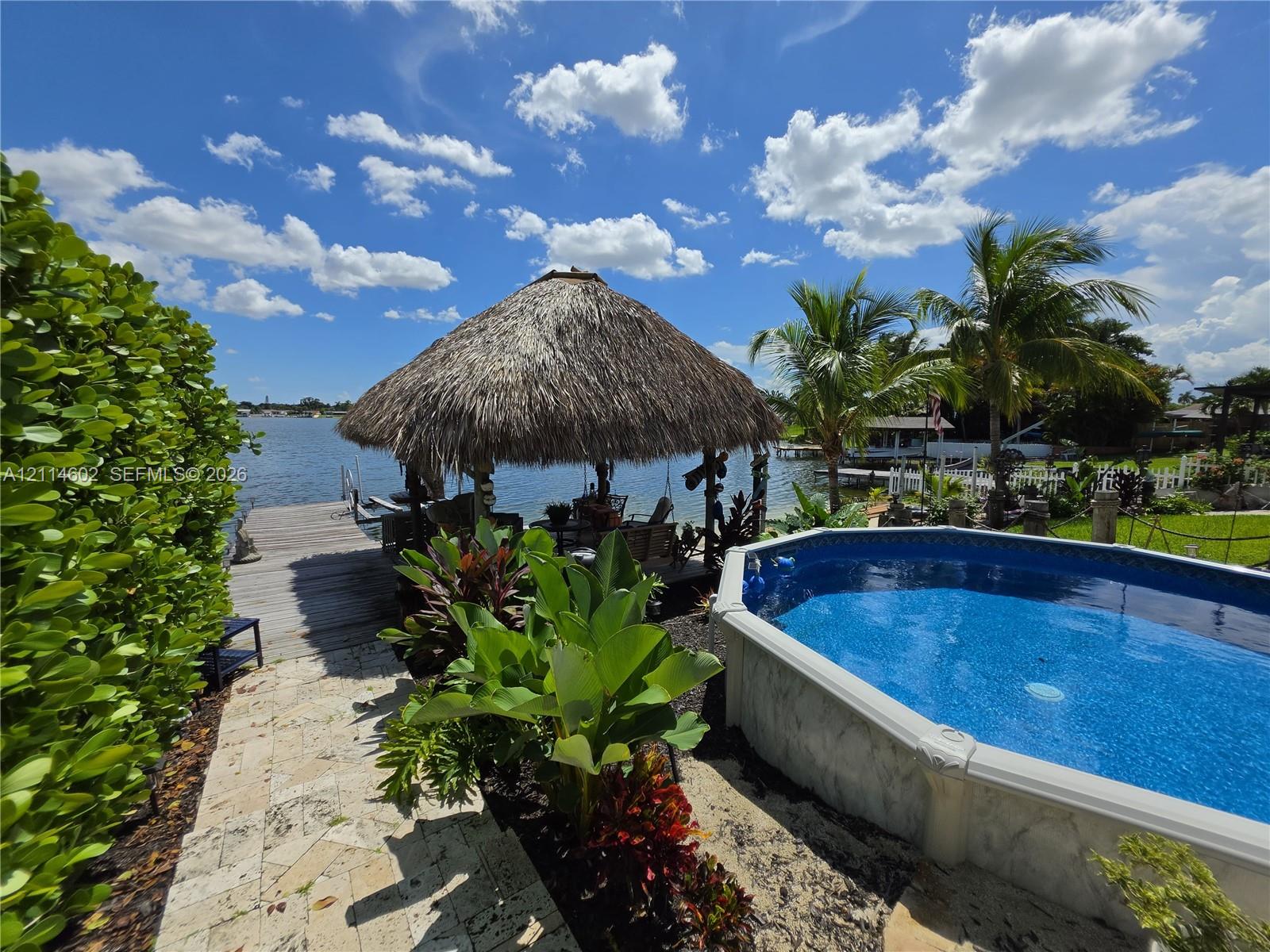 Waterfront backyard featuring an above-ground pool, a thatched-roof tiki hut, a wood-plank dock, and a natural stone-paver walkway