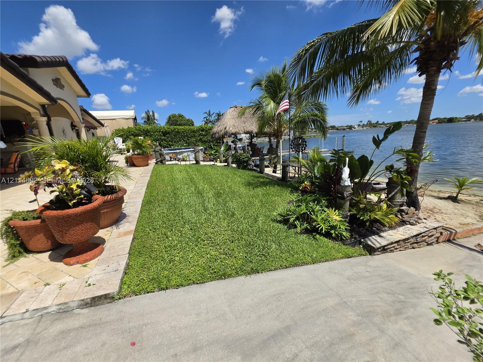 Waterfront backyard featuring a manicured lawn, stone-paved patio, mature palm trees, and a thatched-roof structure