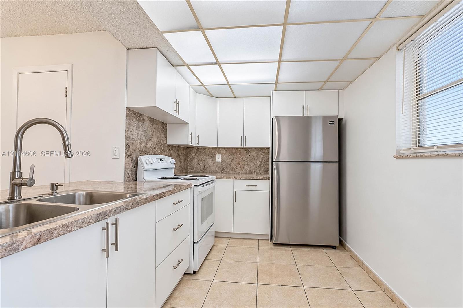 Kitchen featuring white cabinetry, stone-finish countertops, stainless steel refrigerator, tile flooring, and a drop-in sink with a high-arc faucet