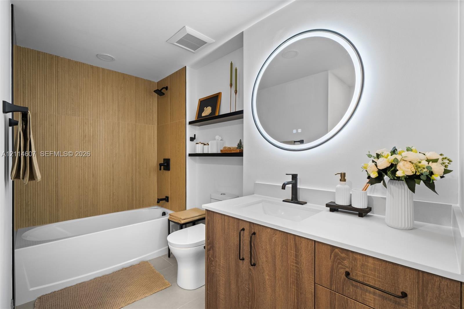 Bathroom featuring a vanity with wood-finish cabinetry, quartz countertop, and integrated sink, illuminated round LED mirror, and a tub with wood-finish tile surround and matte black fixtures