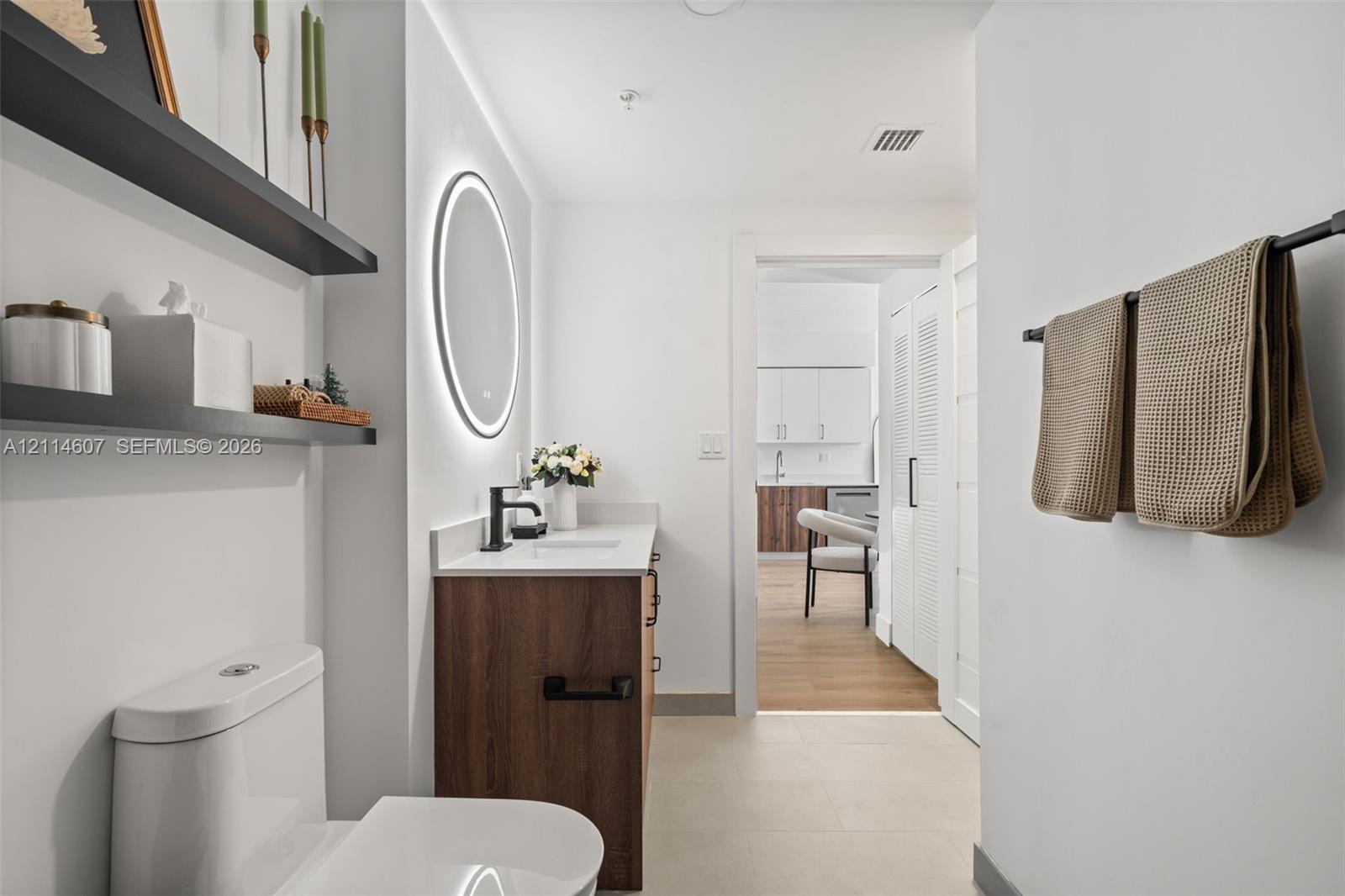 Bathroom featuring a wood-finish vanity with a stone countertop, an integrated sink, a backlit circular mirror, matte black fixtures, and built-in wall shelving