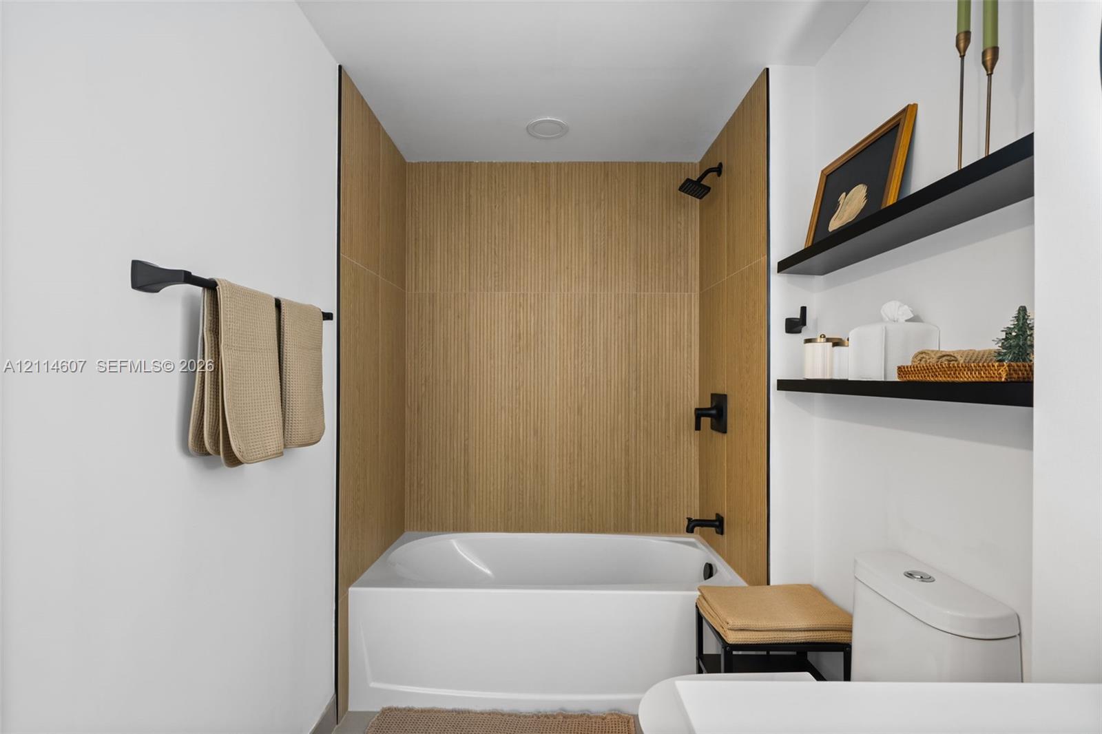 Bathtub enclosure featuring light wood-finish tile surround, matte black plumbing fixtures, and two wall-mounted floating shelves
