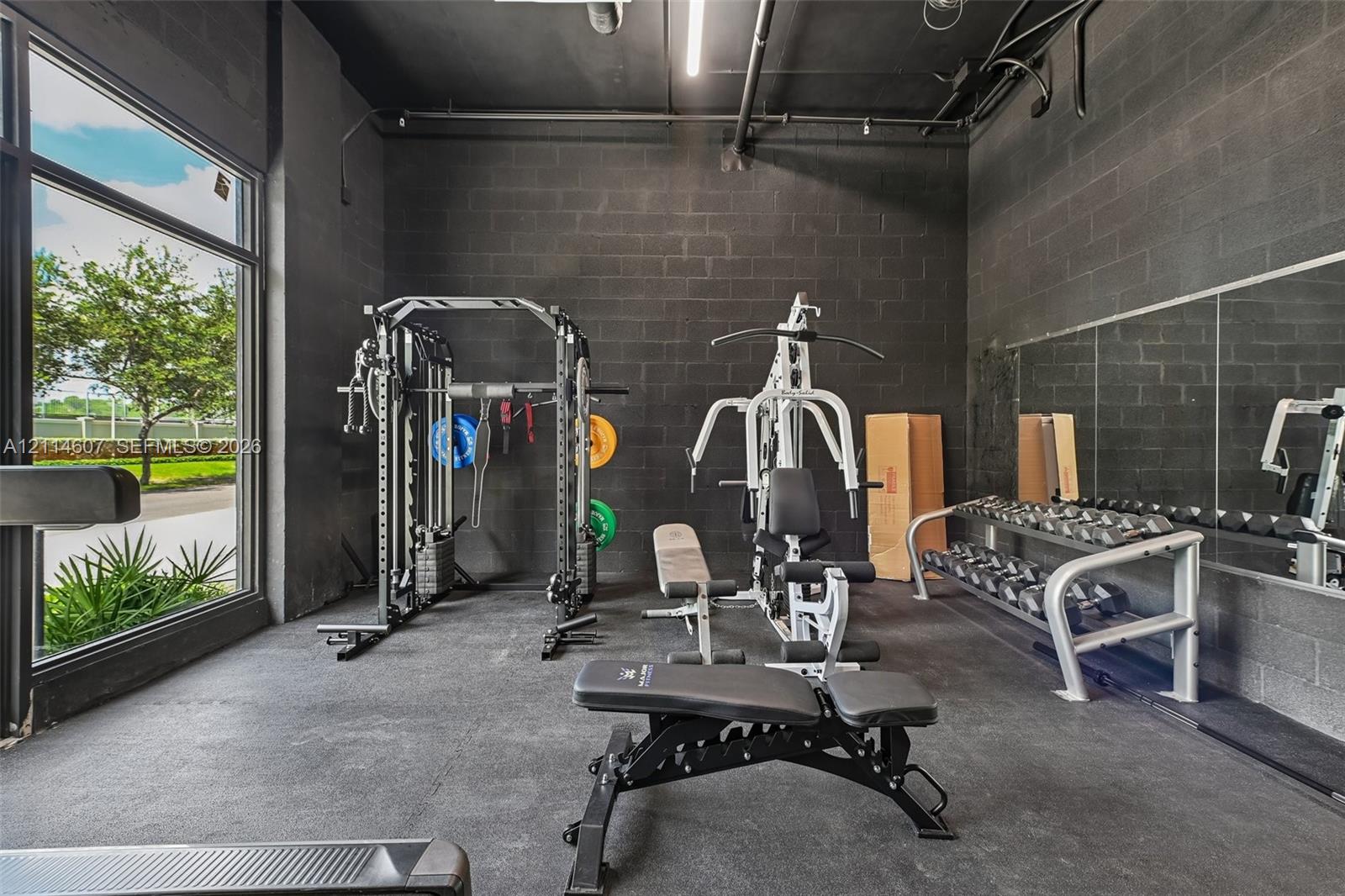 Industrial-style interior space featuring exposed dark-painted concrete block walls, high ceilings with exposed mechanical conduits, floor-to-ceiling windows, and wall-to-wall mirrors