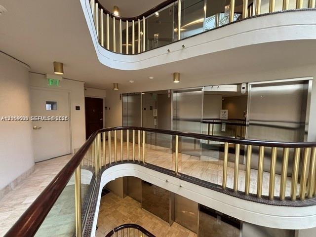 Multi-level atrium featuring a polished stone floor, curved mezzanine with a wood-capped metal railing, and stainless steel elevator doors
