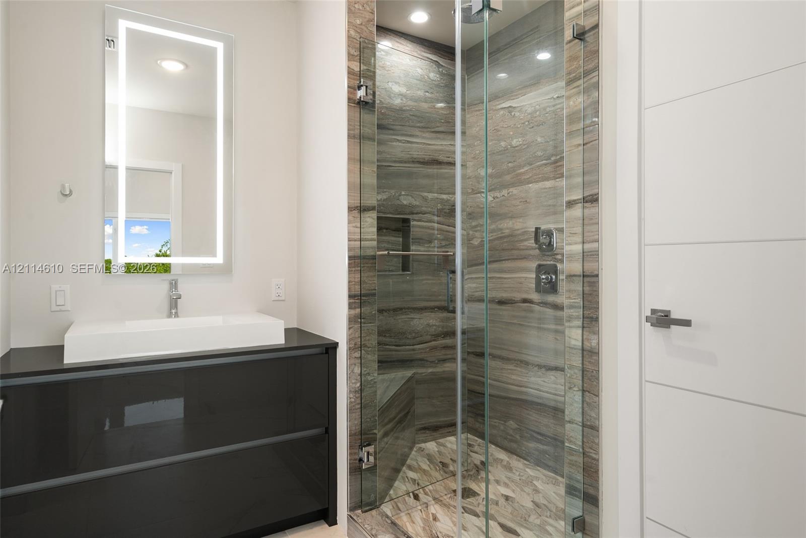 Floating dark-finish vanity with rectangular vessel sink, LED-integrated mirror, walk-in shower with floor-to-ceiling stone-patterned tile, glass shower enclosure, and built-in shower bench