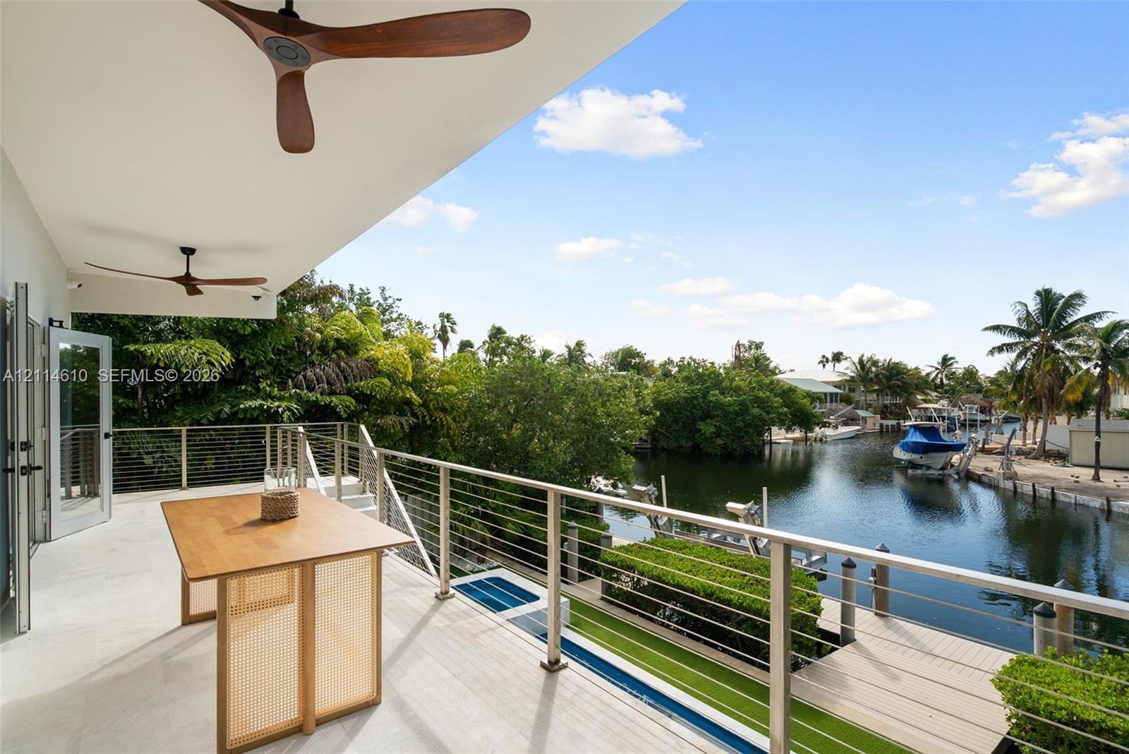 Covered balcony featuring wood-finish ceiling fans, stainless steel cable railing, and light-toned hard flooring, overlooking a canal with a private dock
