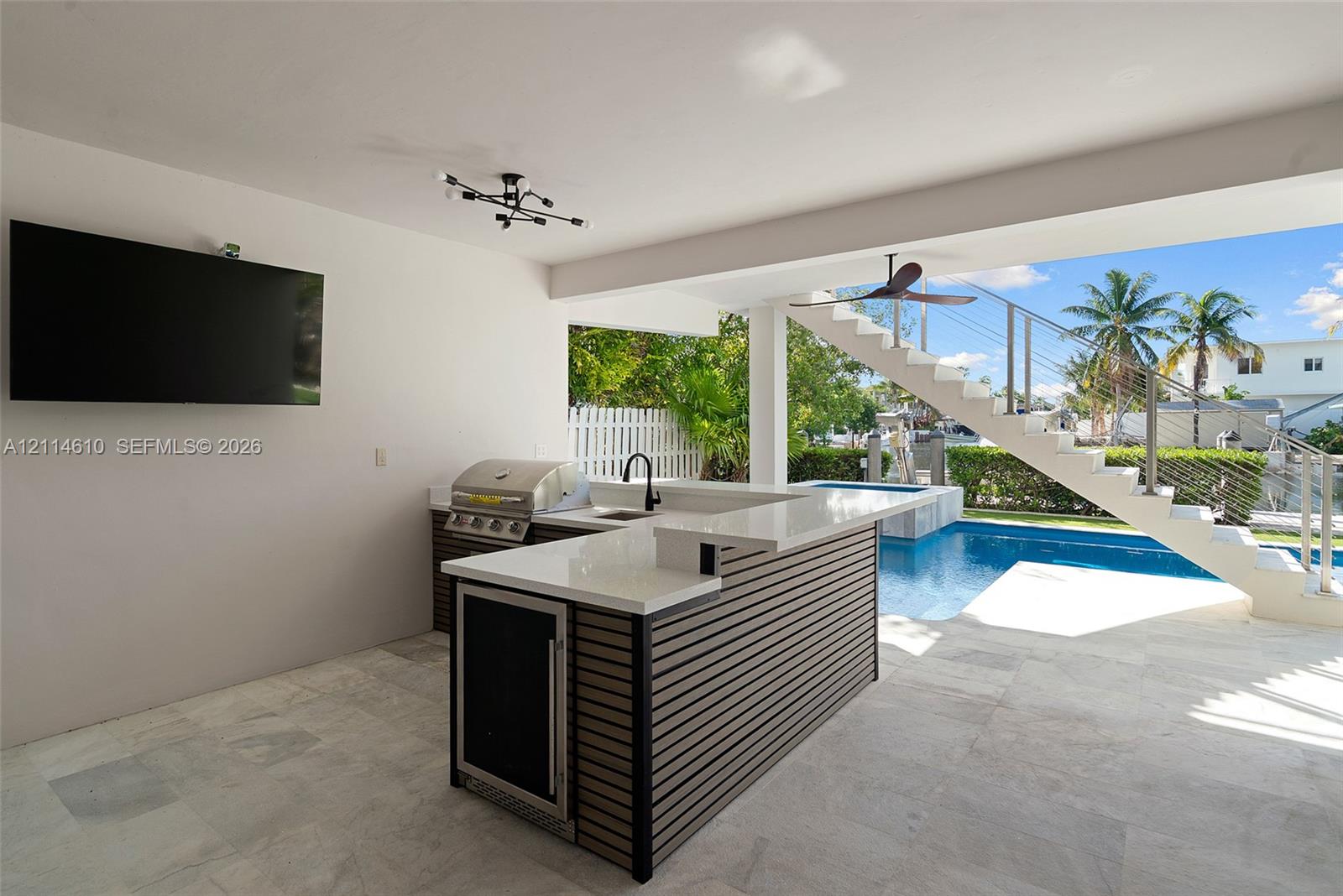 Covered outdoor kitchen featuring a stone-topped island with built-in stainless steel appliances, sleek slatted base cabinetry, stone tile flooring, and integrated ceiling lighting, situated poolside with access to a concrete staircase