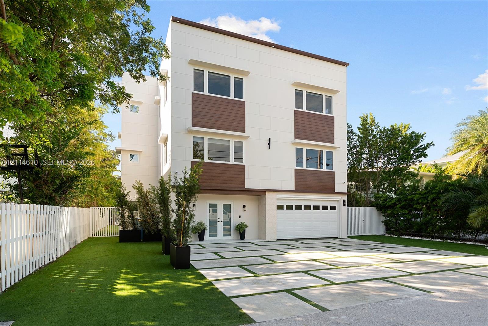 Modern three-story exterior featuring smooth white paneling, horizontal wood-finish siding, a multi-paneled garage door, and a geometric concrete paver driveway with turf accents Modern three-story exterior featuring smooth white paneling, horizontal wood-finish siding, a multi-paneled garage door, and a geometric concrete paver driveway with turf accents