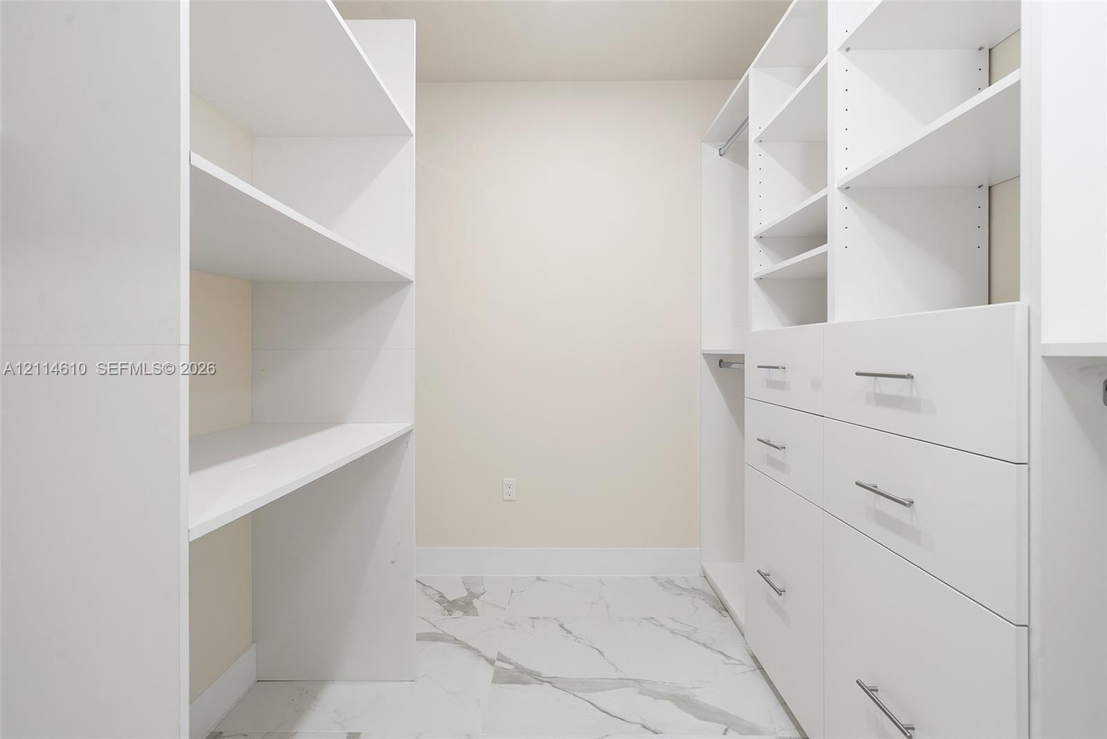 Walk-in closet featuring white custom built-in shelving, integrated hanging rods, and multi-drawer cabinetry with metallic pulls finished in polished chrome, complemented by large-format stone-pattern tile flooring