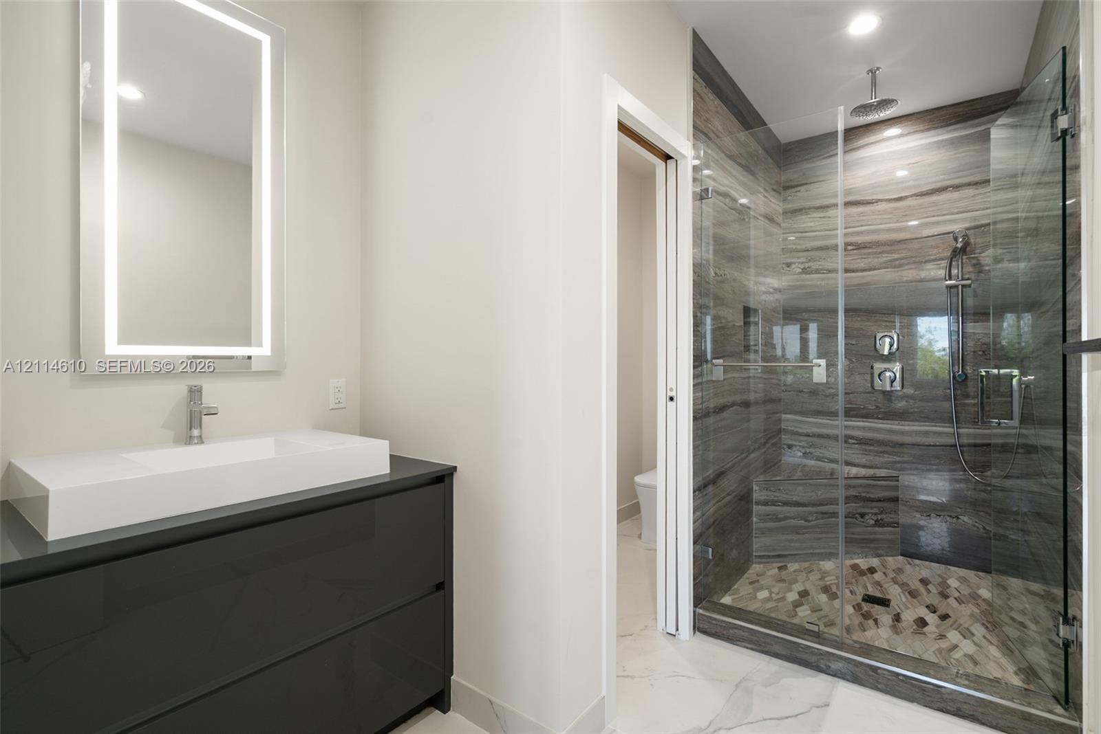 Modern bathroom featuring a dark floating vanity with an integrated rectangular sink, wall-mounted LED-backlit mirror, and a walk-in shower with floor-to-ceiling stone-patterned tile, a rainfall showerhead, and a glass enclosure