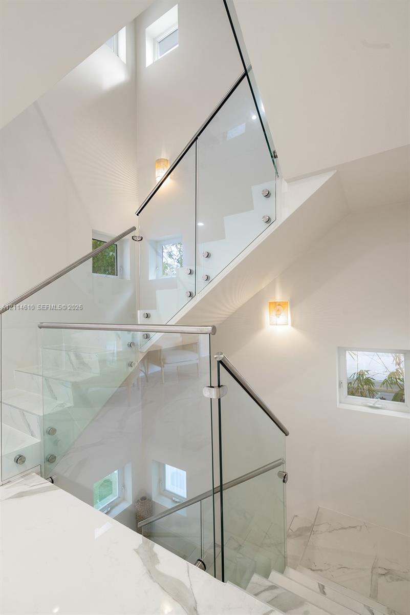 Staircase featuring white stone treads, frameless glass railings with metal standoffs, stainless steel handrails, multiple square windows, and wall-mounted light fixtures