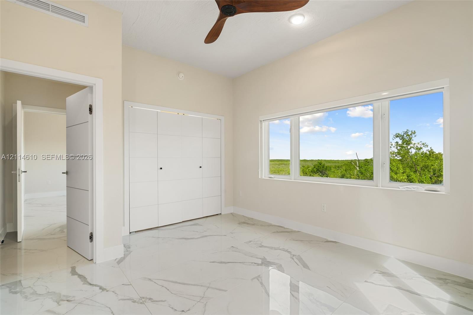Interior space featuring large-format tile flooring with stone-style veining, triple-panel built-in closet, multi-pane window with exterior natural scenery, and a ceiling-mounted fan with wood-finish blades