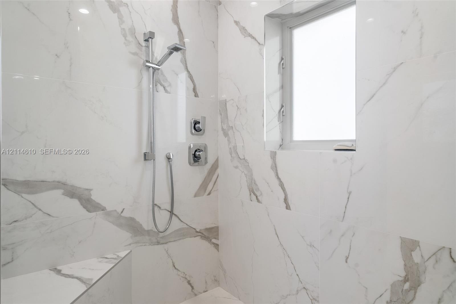 Walk-in shower featuring large-format veined tile surround, polished chrome adjustable handheld shower fixture, integrated tiled bench, and a rectangular transom window