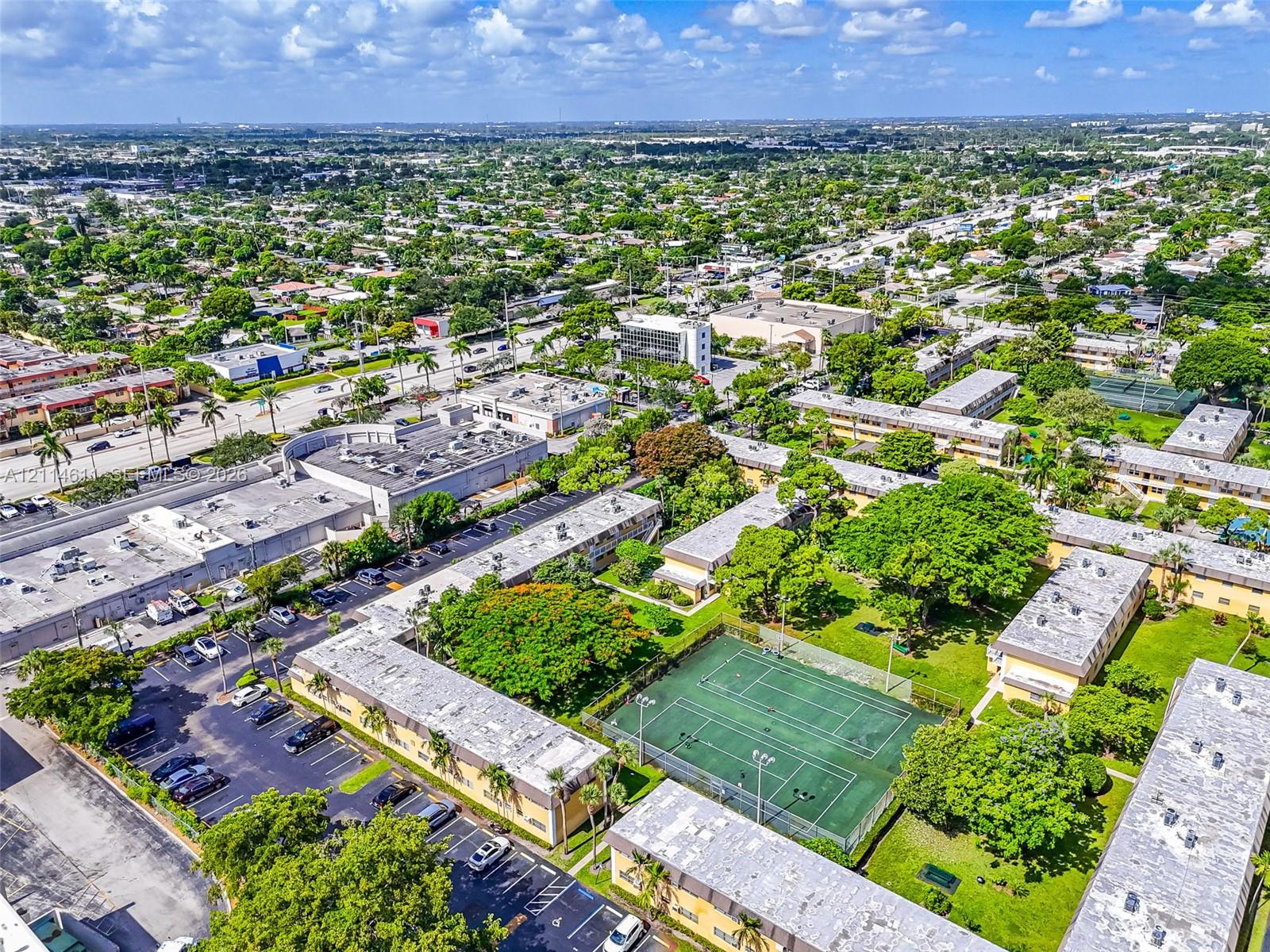 Aerial perspective of the residential complex featuring a fenced tennis court, paved surface parking, expansive lawn areas, and mature trees