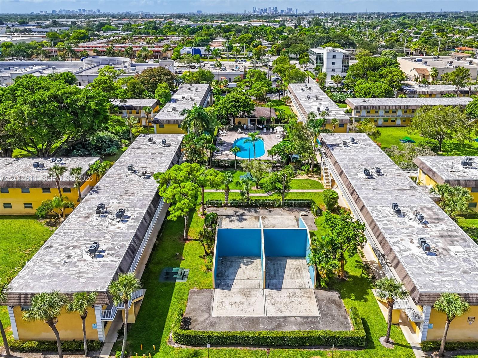 Aerial perspective of a multi-building property featuring a central outdoor swimming pool, dedicated racquetball courts, extensive manicured lawn areas, and mature palm trees