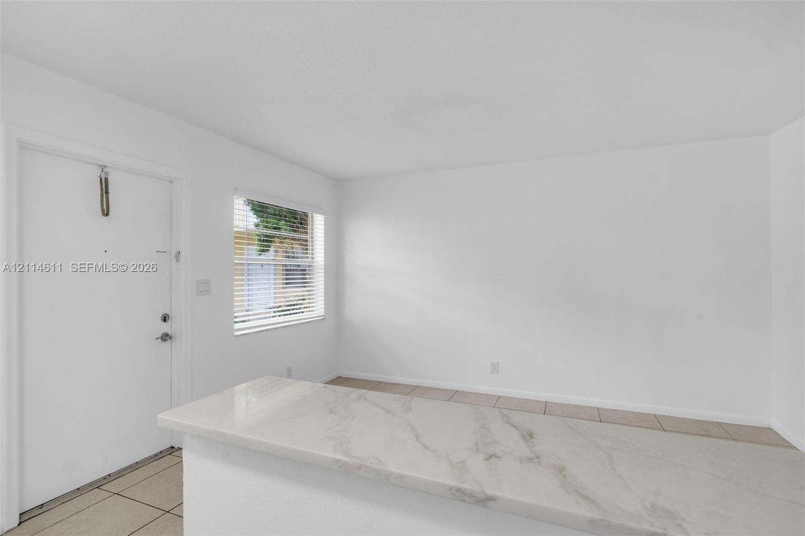 Interior space featuring white walls, neutral tile flooring, horizontal window blinds, and a countertop with a marble-patterned finish Interior space featuring white walls, neutral tile flooring, horizontal window blinds, and a countertop with a marble-patterned finish