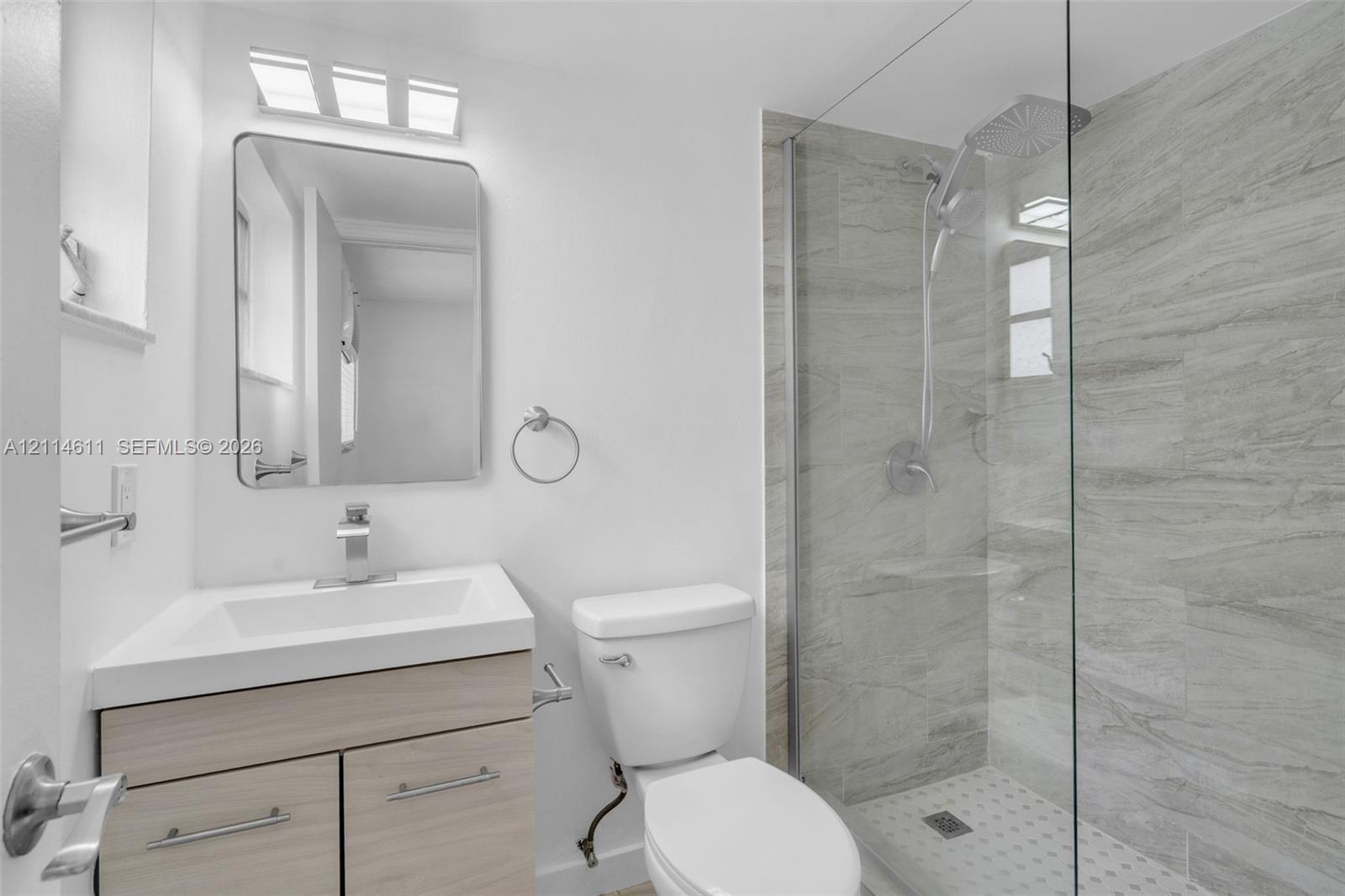 Modern vanity with a rectangular sink, wood-finish cabinetry, frameless glass walk-in shower with stone-tile surround, and rainfall shower head