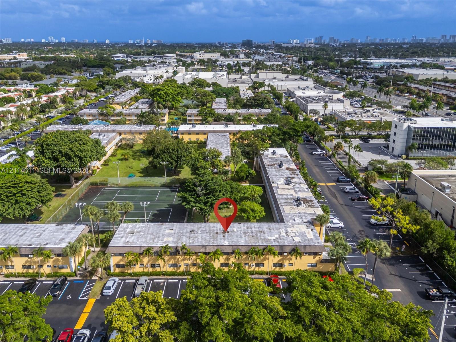 Aerial view of residential property featuring a tennis court, manicured grounds, mature trees, and ample paved surface parking Aerial view of residential property featuring a tennis court, manicured grounds, mature trees, and ample paved surface parking