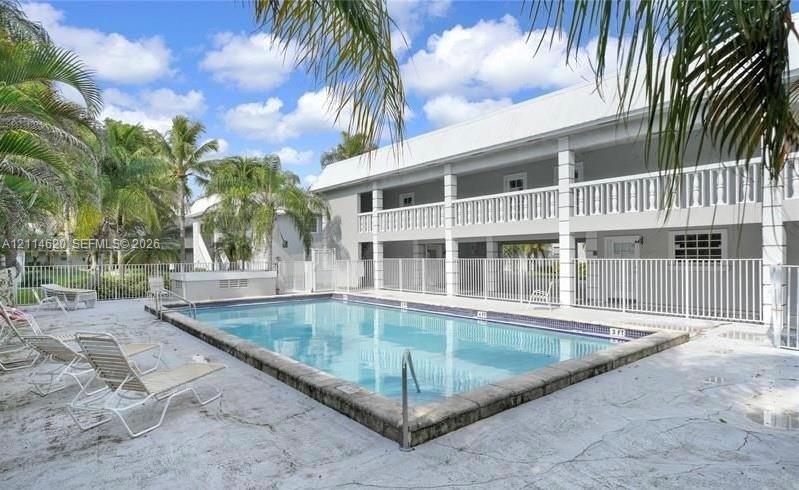 In-ground swimming pool with stone-style coping, enclosed by a white metal safety fence, featuring a two-story building exterior with a white balustrade balcony and palm-lined grounds