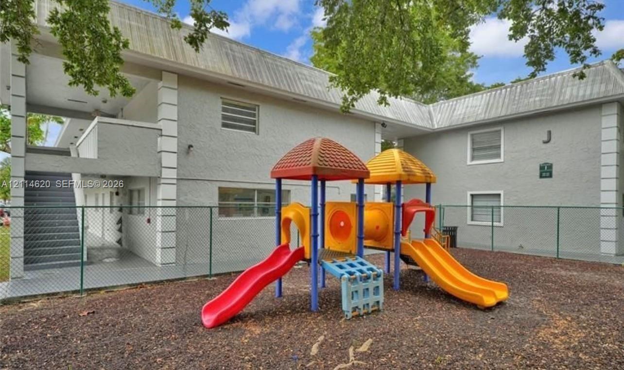 Multi-story stucco exterior building featuring a fenced-in recreation area with a modular play structure and wood-chip ground cover