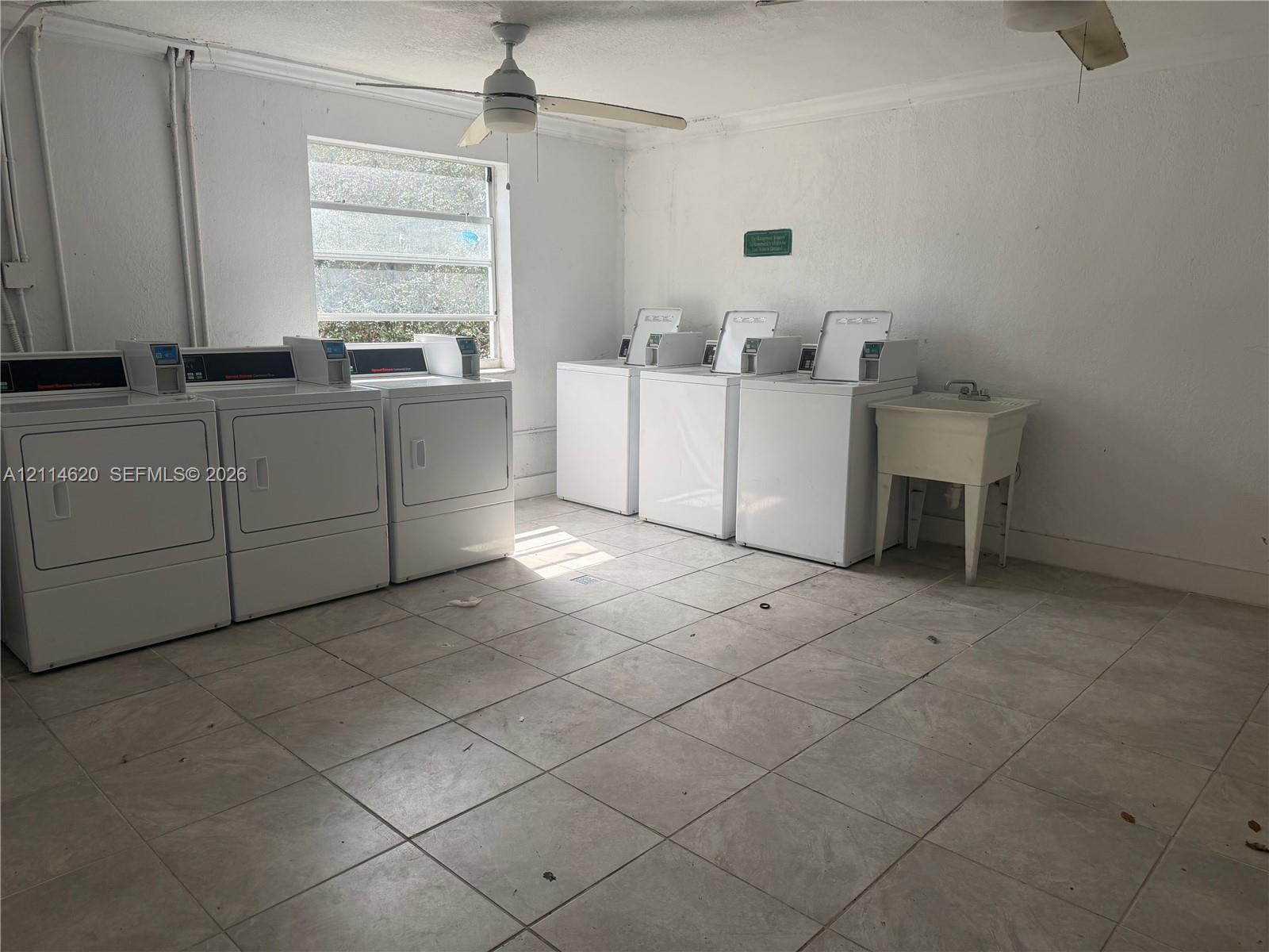 Utility room featuring ceramic tile flooring, three large-capacity washing machines, three matching clothes dryers, and a built-in utility sink