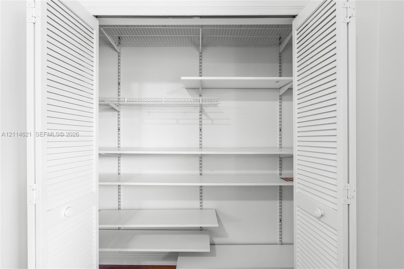 Storage closet featuring white louvered bifold doors, adjustable shelving tracks, and multiple solid surface shelves