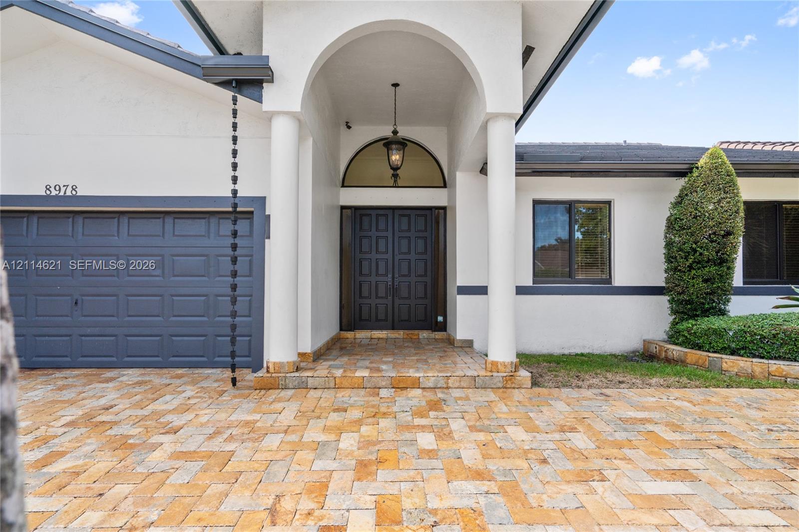 Exterior facade featuring a herringbone-patterned paver driveway, arched entryway with dual columns, double front doors with a semi-circular transom window, and a metal chain-link downspout