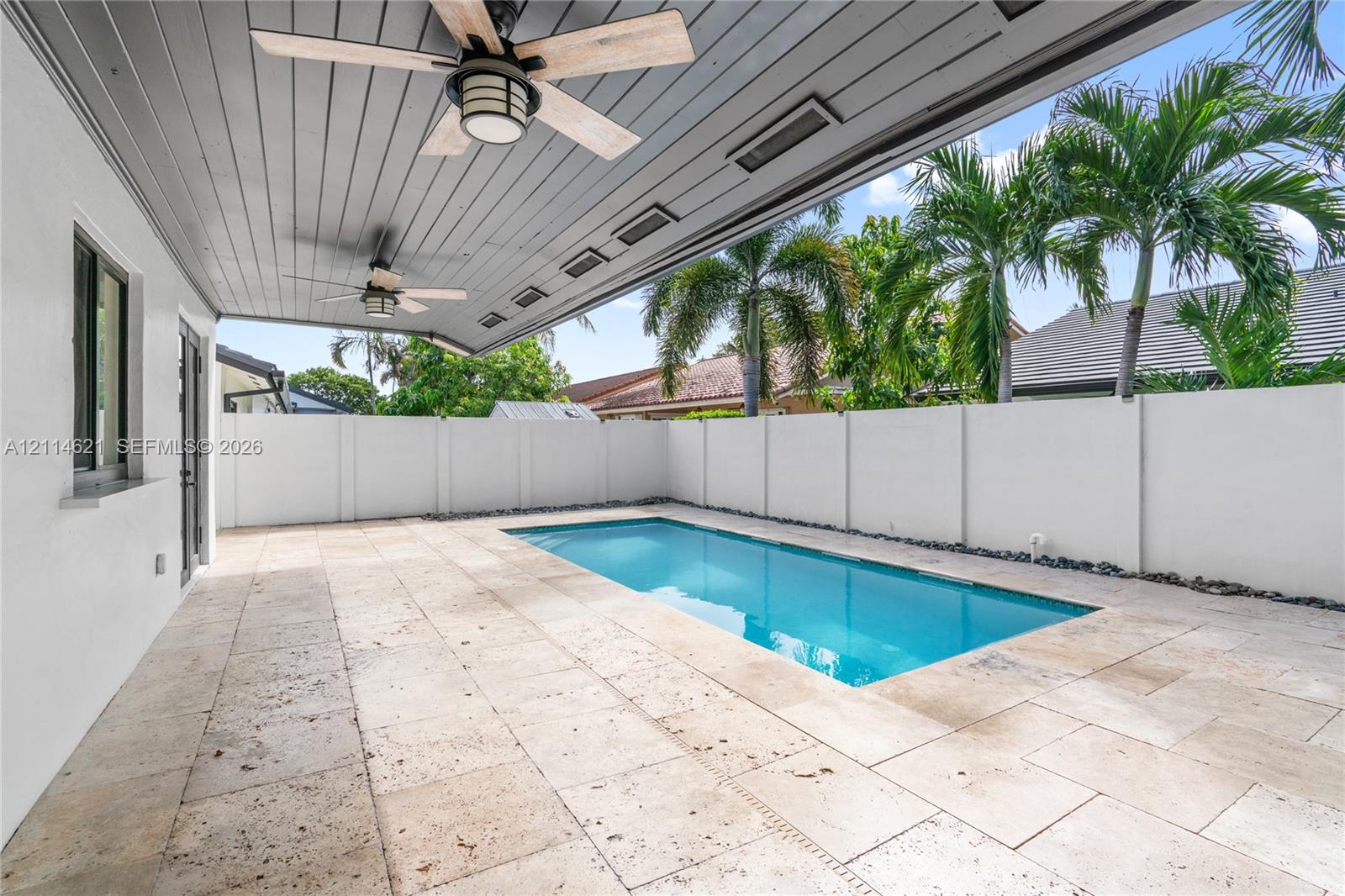 Covered patio featuring a grey tongue-and-groove ceiling with dual ceiling fans, stone paver decking, and a rectangular swimming pool enclosed by a white privacy wall