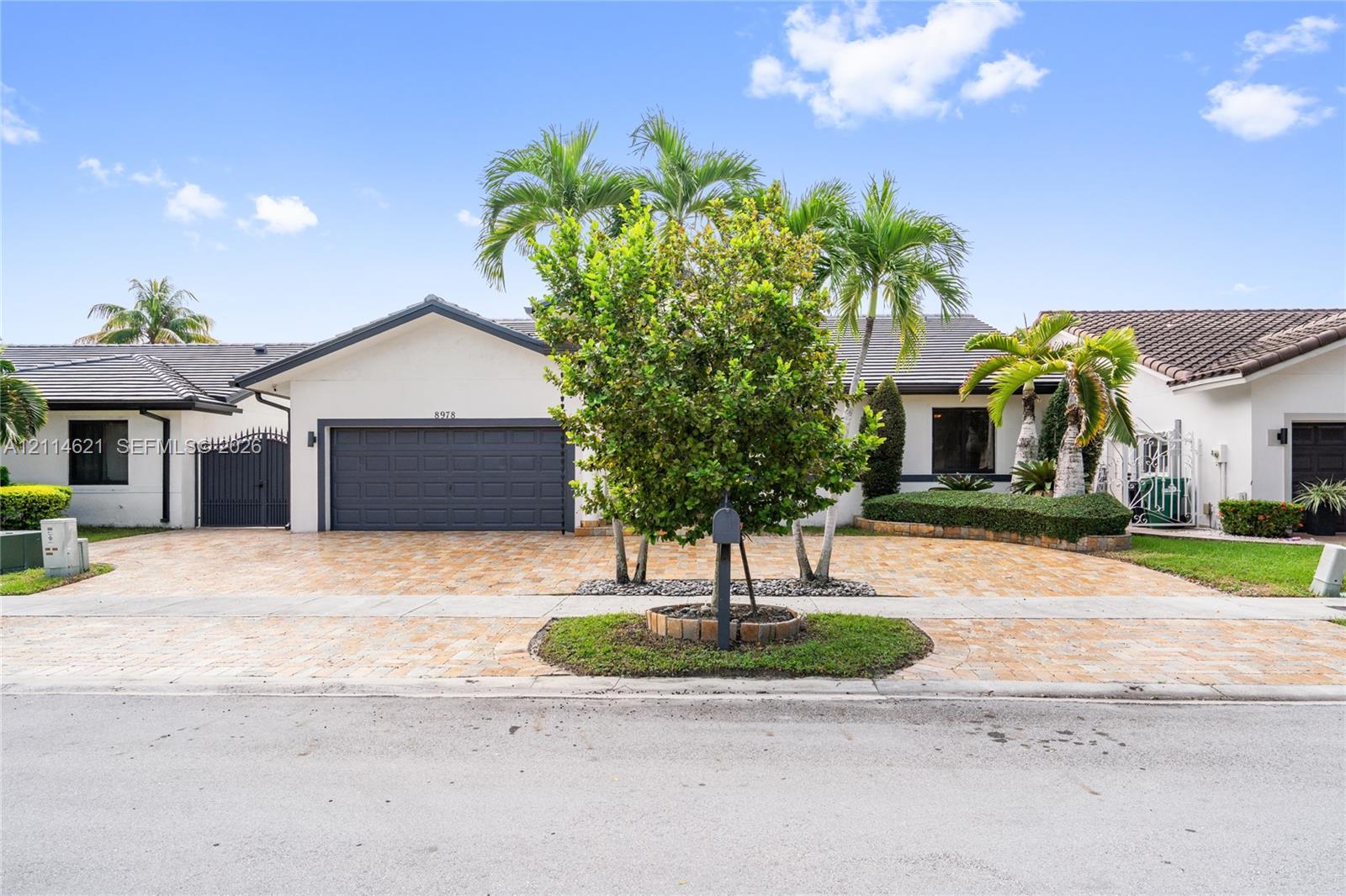 Single-story exterior featuring a light-toned stucco faade, a multi-car garage with a paneled door, a paved driveway, a tile roof, and curated tropical landscaping