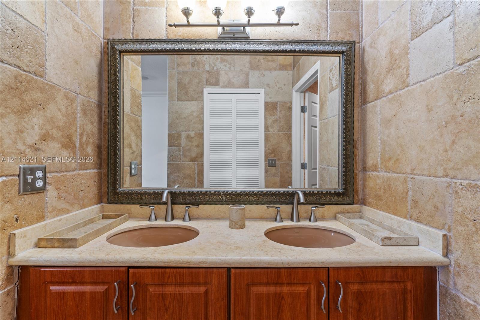 Dual-sink vanity with a stone countertop, wood cabinetry, stone tile wall cladding, and a large framed mirror