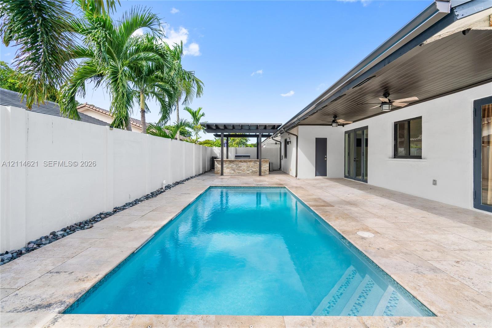 Outdoor space featuring a rectangular swimming pool, stone-tiled patio, built-in outdoor bar with a pergola, covered lanai with ceiling fans, and a solid perimeter privacy wall Outdoor space featuring a rectangular swimming pool, stone-tiled patio, built-in outdoor bar with a pergola, covered lanai with ceiling fans, and a solid perimeter privacy wall