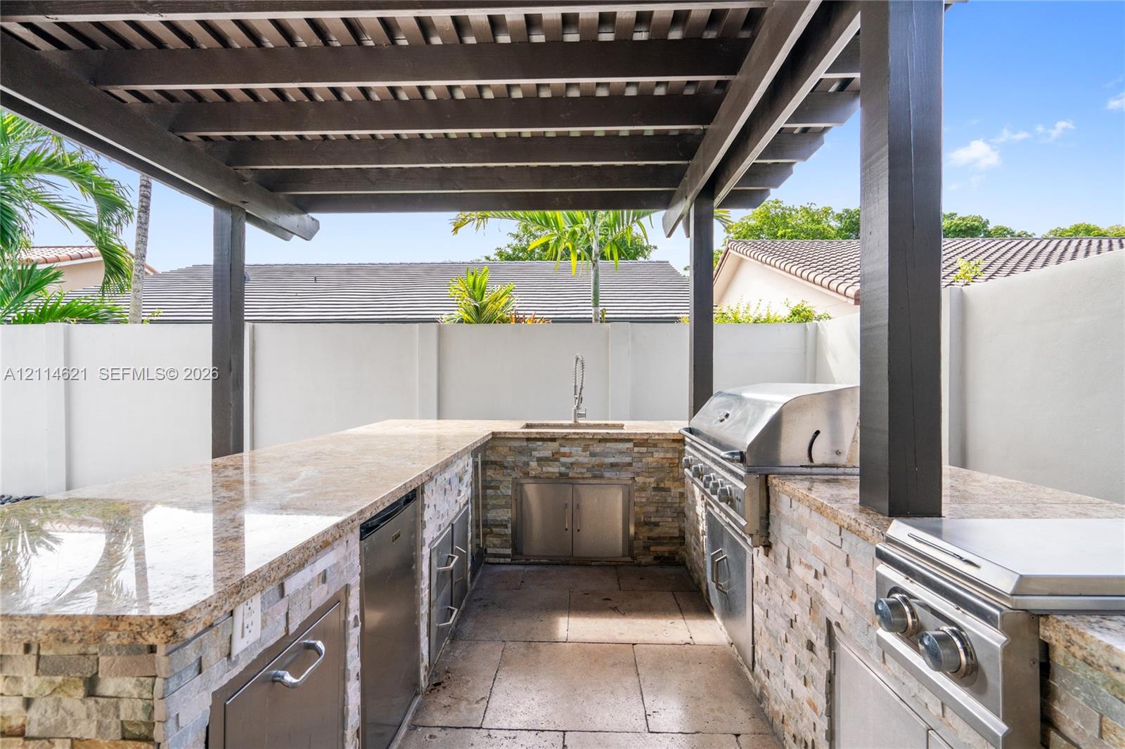 Outdoor kitchen featuring a stone-clad base, built-in stainless steel grill, stone countertop, undermount sink, and overhead pergola Outdoor kitchen featuring a stone-clad base, built-in stainless steel grill, stone countertop, undermount sink, and overhead pergola