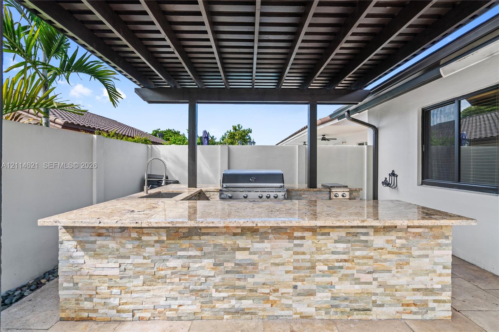 Outdoor kitchen featuring a stacked-stone base, natural stone countertop, built-in stainless steel grill, integrated sink, and a covered pergola structure