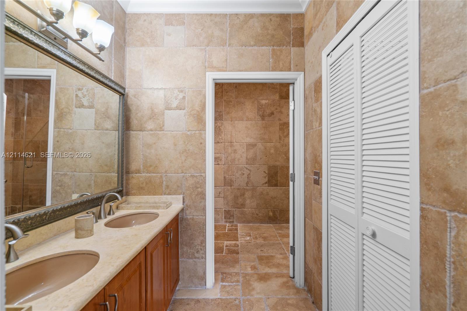 Natural stone tile wall and floor surfaces, dual-basin vanity with a stone countertop, framed wall mirror, and louvered-style closet doors