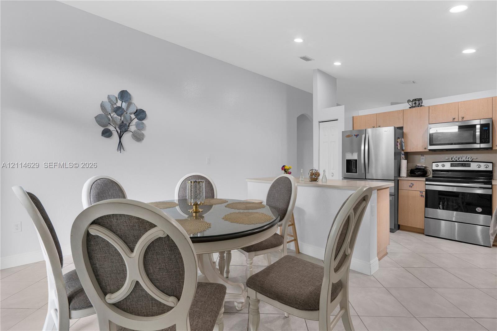Kitchen featuring light wood-finish cabinetry, stainless steel appliances, and a breakfast bar with a neutral countertop, complemented by neutral tile flooring and recessed lighting