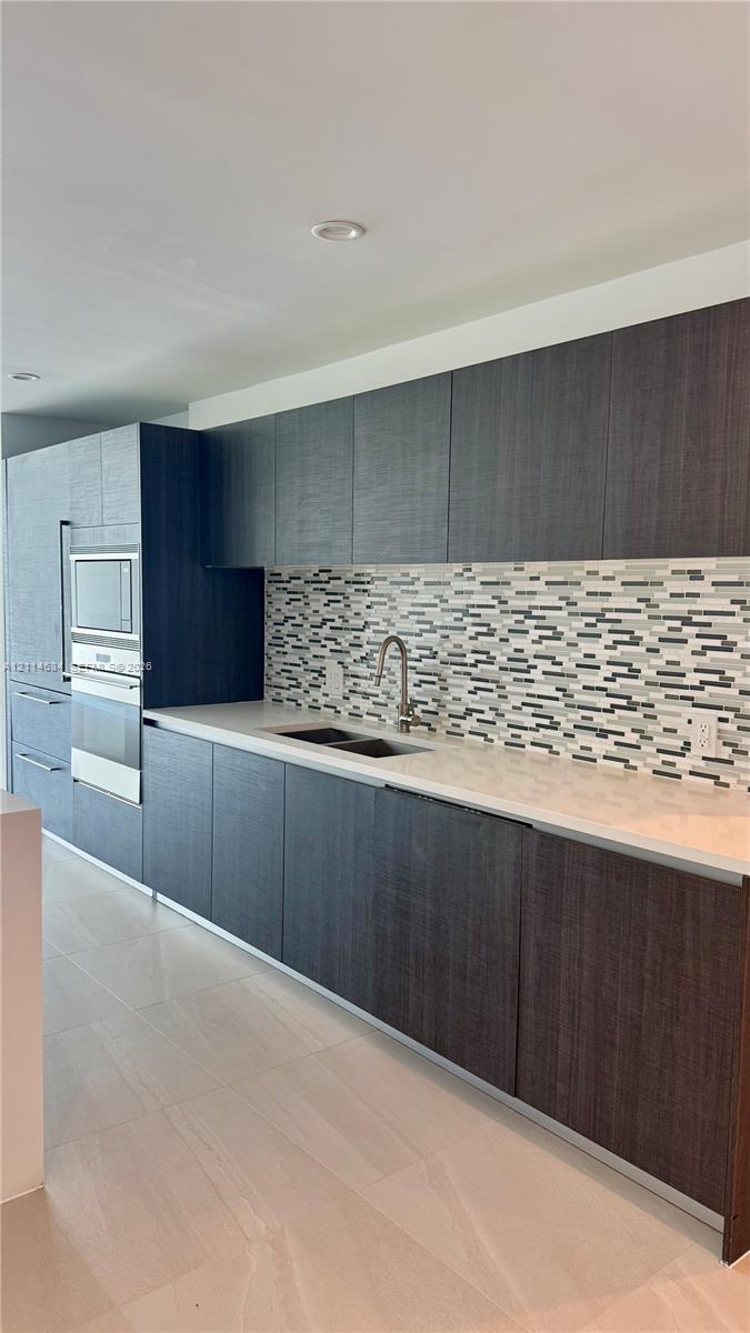 Dark-toned cabinetry featuring flat-panel doors, stone-surface countertops, mosaic tile backsplash, integrated stainless steel oven, and recessed ceiling lighting
