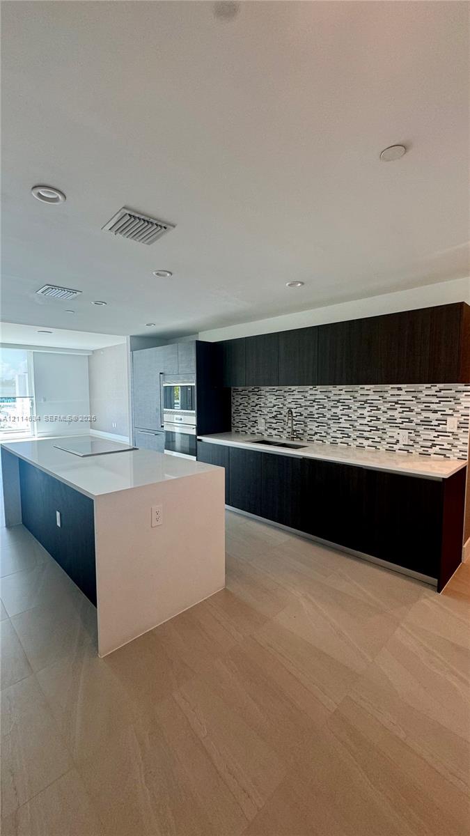 Kitchen featuring dark-toned flat-panel cabinetry, quartz countertops, a stone mosaic backsplash, a large center island, and recessed lighting