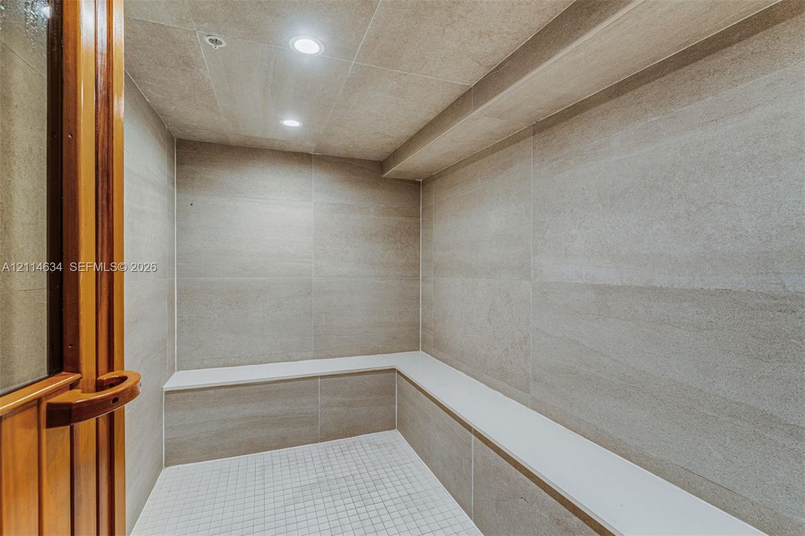 Steam room featuring large-format stone-tile walls, built-in tiered stone seating, mosaic-tile flooring, and recessed lighting behind a wood-framed glass door