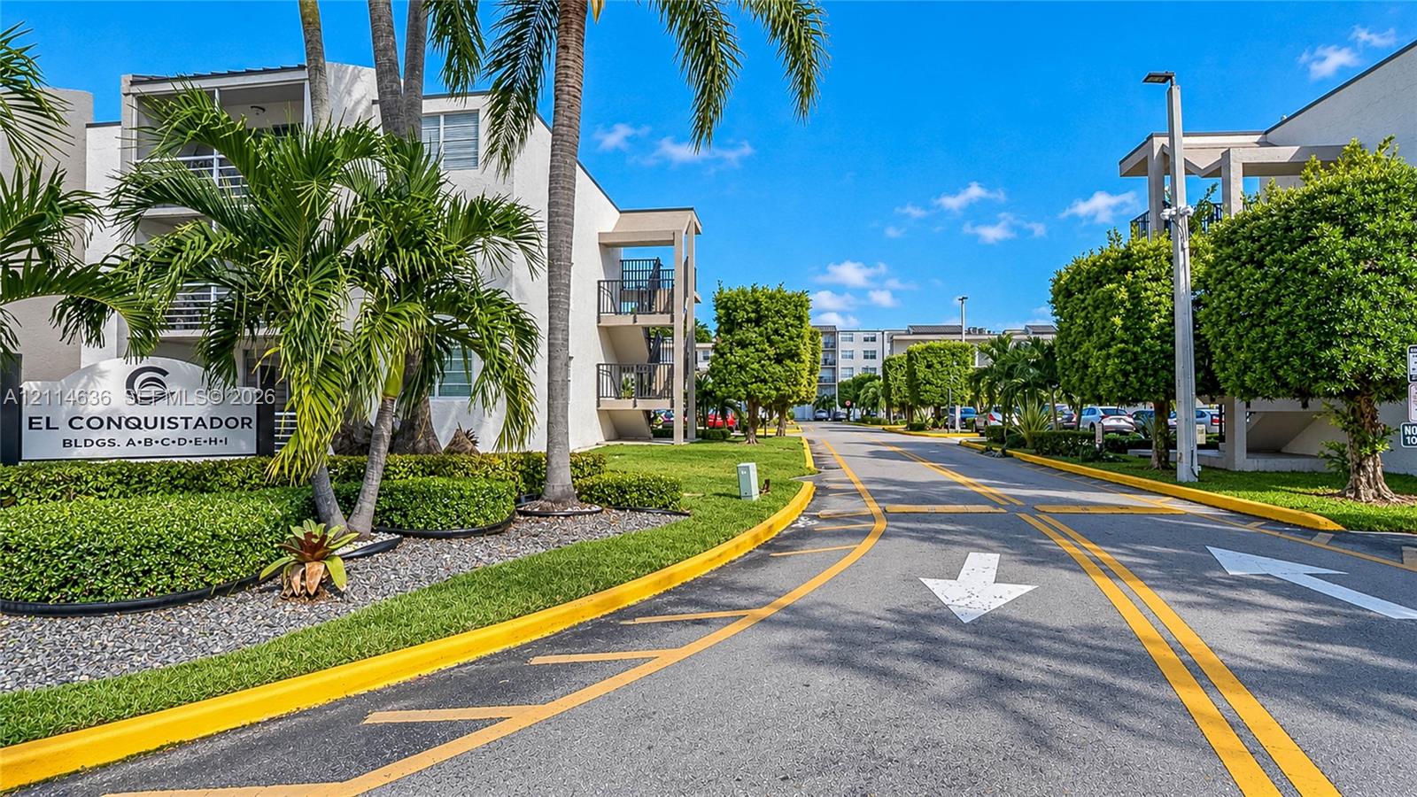Exterior entrance featuring stucco-finished buildings, mature palm trees, manicured hedging, and paved roadway with directional pavement markings