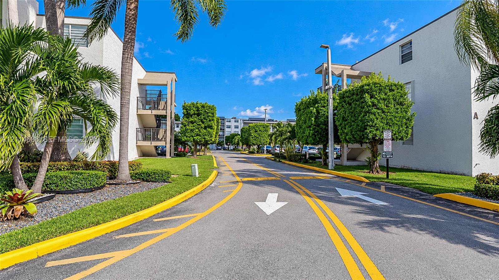 Multi-story exterior featuring a light-toned stucco facade, paved driveways, manicured hedges, and mature palm trees