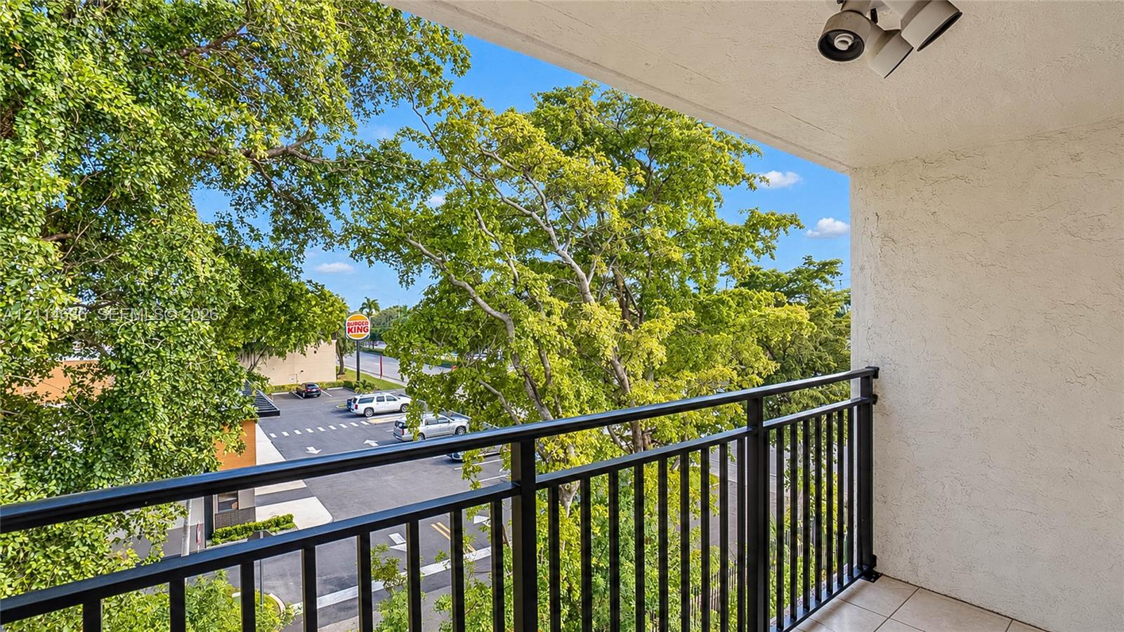 Covered balcony featuring textured stucco walls, tile flooring, a black metal railing, and overhead lighting