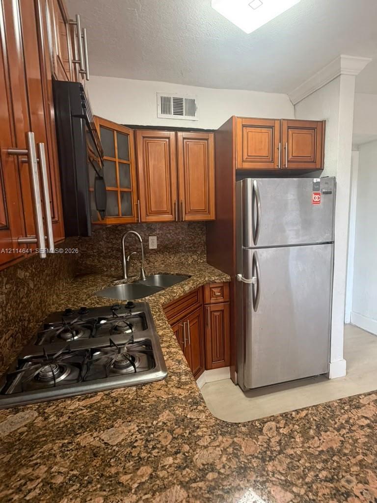Kitchen featuring wood cabinetry, stone countertops, stainless steel refrigerator, integrated gas cooktop, and a double-basin undermount sink with a high-arch faucet