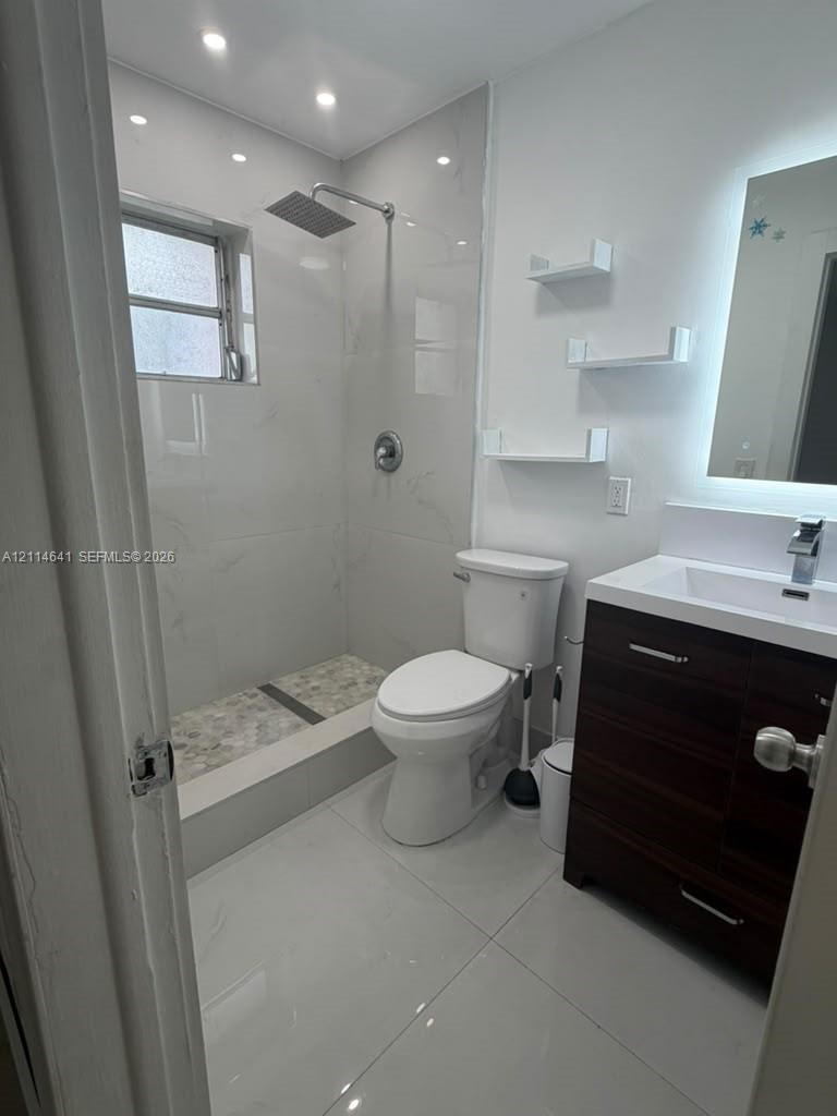 Full bathroom featuring large-format floor tile, walk-in shower with stone-style pebble flooring and rainfall showerhead, floating vanity with integrated countertop, and LED-backlit mirror