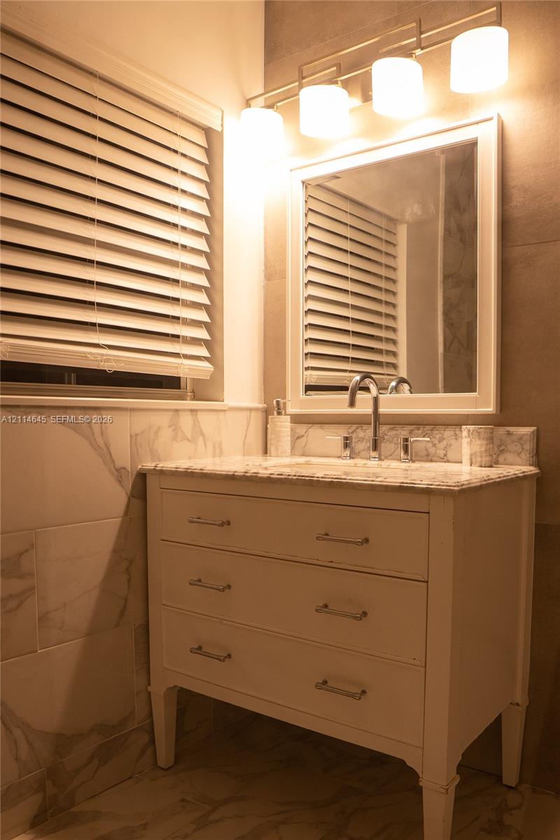 Bathroom vanity featuring a stone countertop, white cabinetry with polished chrome pulls, a framed wall mirror, a four-light fixture, and large-format wall tile
