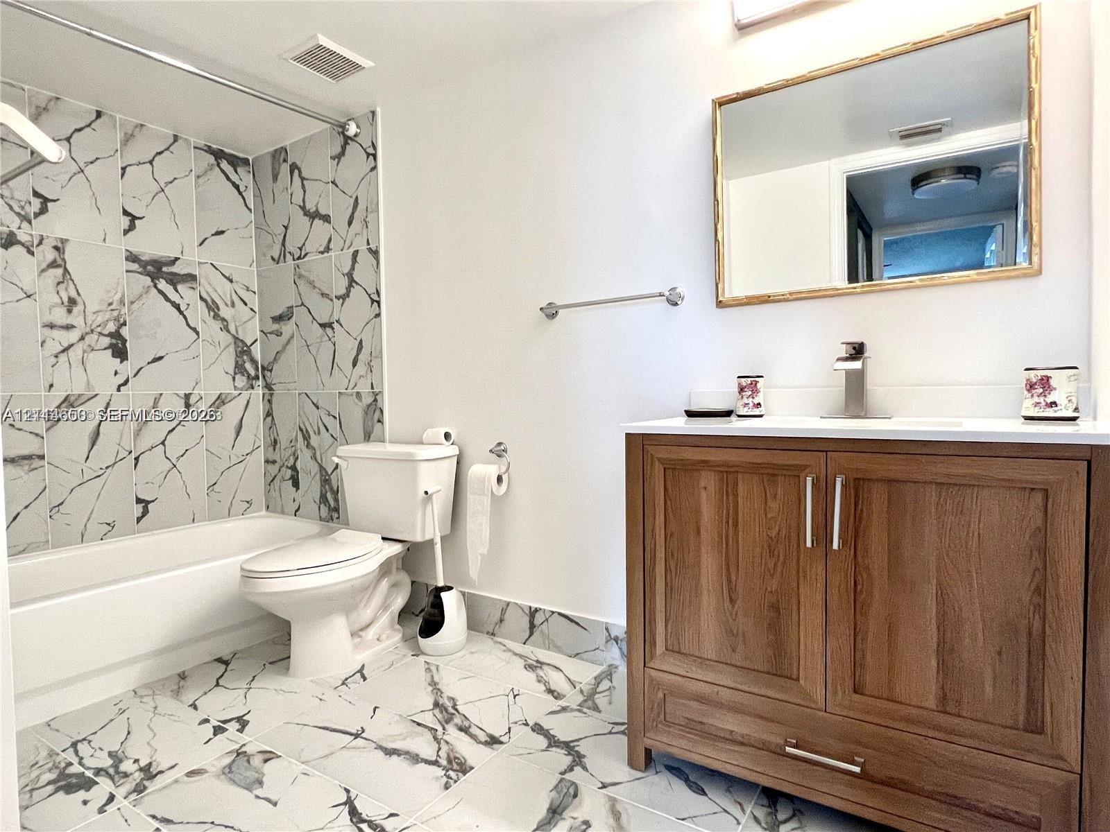 Bathroom featuring large-format stone-patterned tile flooring, a wood-finish vanity with a white countertop, wall-mounted mirror, and a tub-shower surround accented with coordinating stone-patterned wall tile